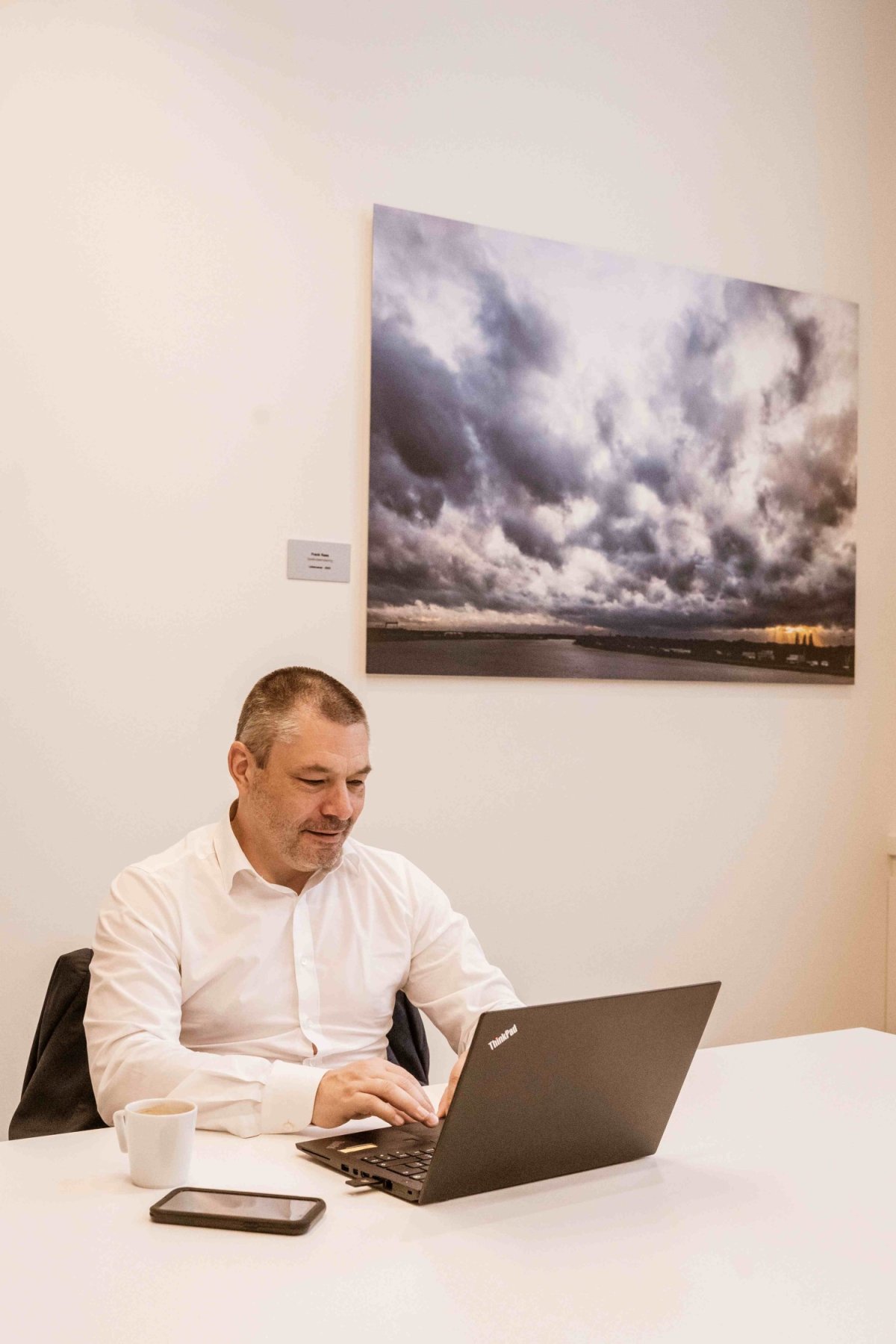 Een man in een wit overhemd zit aan een bureau met een laptop, een smartphone en een kopje koffie, terwijl hij op de laptop typt. Op de muur achter hem hangt een kunstwerk met een afbeelding van bewolkte lucht en een landschap.