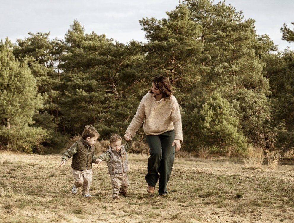 Een vrouw wandelt hand in hand met twee jonge kinderen in een bosrijke omgeving.