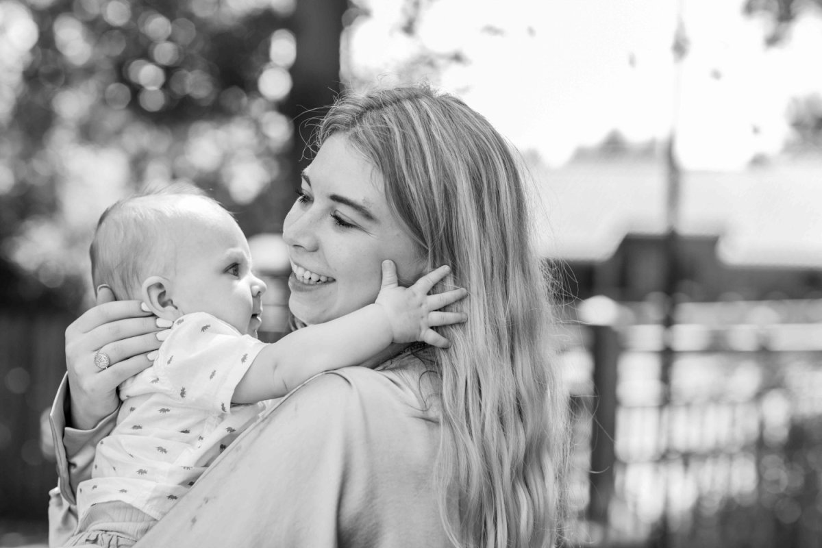 Een oudere vrouw met grijzend haar en bril omarmt een lachend kindje met blond haar, terwijl ze samen in een bosrijke omgeving staan.