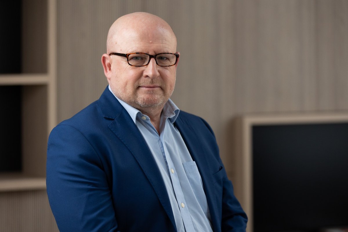 Een professionele man met een bril, gekleed in een blauwe blazer en een lichtblauw overhemd, poseert voor een neutrale achtergrond van houten panelen en een zwart scherm.