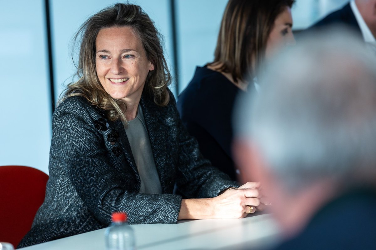 Een vrouw met een glimlach, zittend aan een vergadertafel, gekleed in een grijze blazer, met een achtergrond van andere deelnemers aan de vergadering.