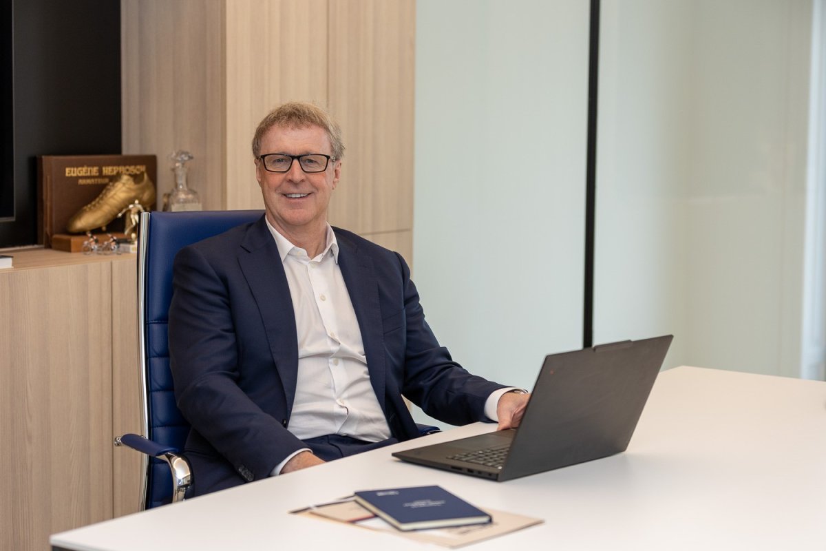 Een man in een blauw pak zit aan een bureau met een laptop, glimlachend naar de camera. Achter hem staan boeken en een gouden schoen op de kast.