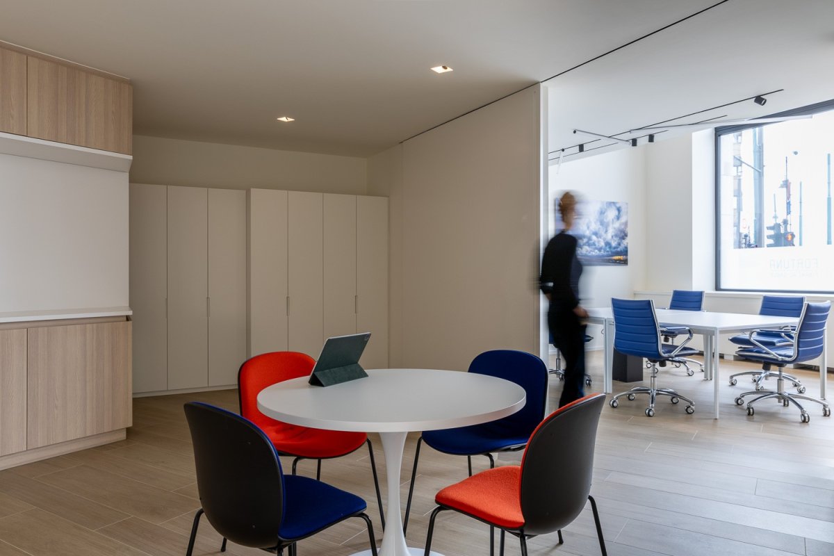 Moderne kantoorruimte met een ronde tafel en kleurrijke stoelen, een persoon in beweging op de achtergrond, en lichtinval van een raam.