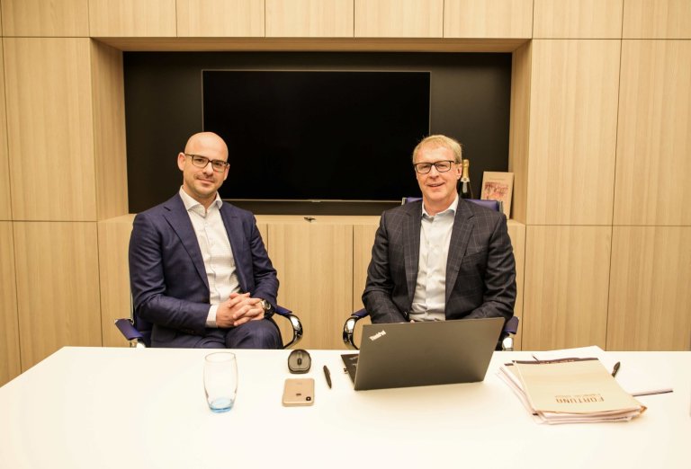 Twee mannen in zakelijke kleding zitten aan een tafel in een kantoor met een laptop en een glas water.