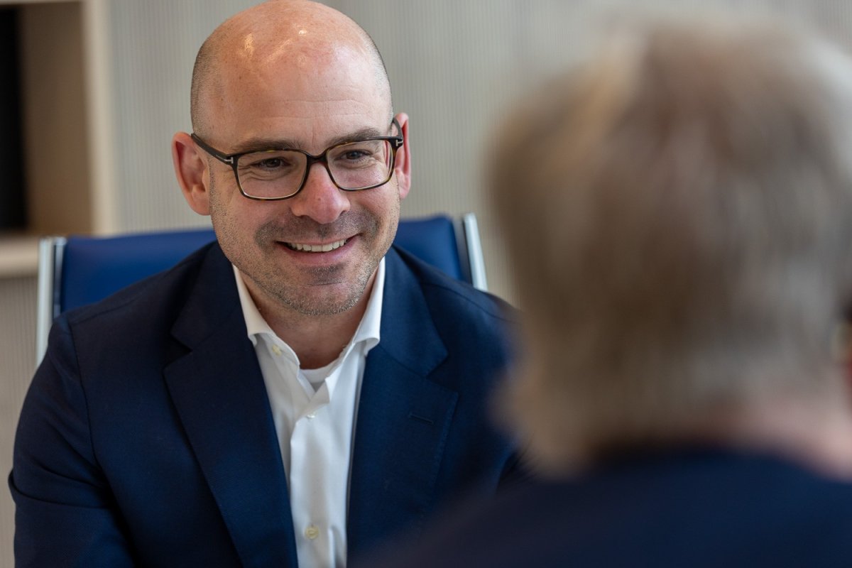 Een man met een bril en een gladgeschoren hoofd glimlacht tijdens een gesprek, terwijl hij tegenover een andere persoon zit.