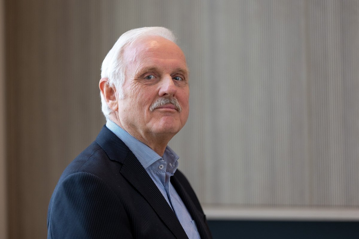 Een oudere man met grijs haar en een snor, gekleed in een donker jasje en een lichtblauw overhemd, kijkt naar de camera tegen een neutrale achtergrond.