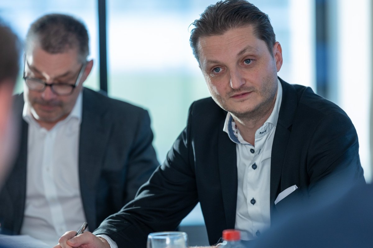 Twee mannen in formele kleding zitten aan een vergadertafel, met de focus op de man rechts die naar de camera kijkt en aandachtig luistert. De man links is iets aan het lezen. Op de tafel staan glazen en flessen.