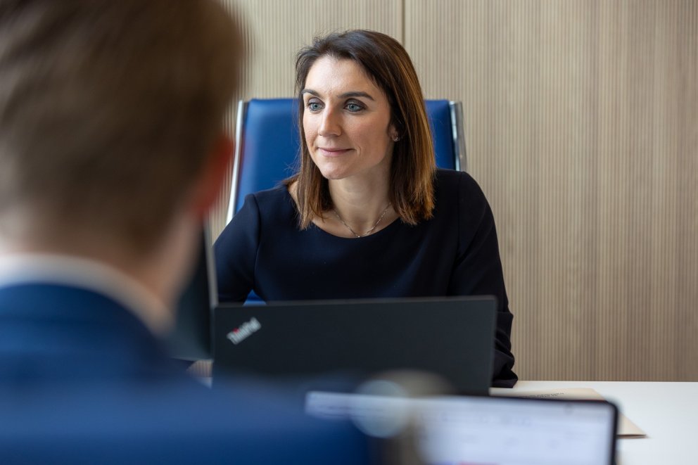 Een vrouw in een zakelijke outfit zit aan een bureau met een laptop, terwijl ze aandachtig naar een persoon tegenover haar kijkt, waarvan alleen het hoofd zichtbaar is.