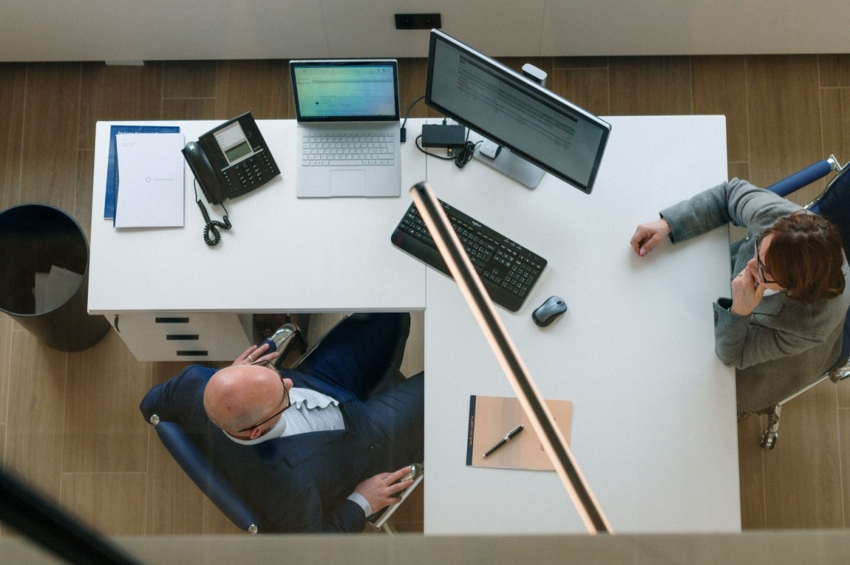 Twee personen zitten aan een bureau in een kantooromgeving, omringd door duidelijk zichtbare technologie zoals laptops, een telefoon en een toetsenbord. Een van hen is aan het bellen, terwijl de ander aandachtig kijkt en nadenkt.