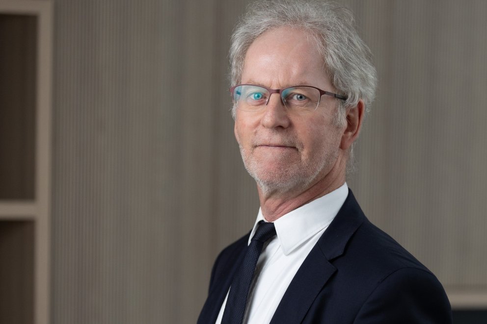Portret van een man met grijs haar en een baard, draagt een donkerblauw pak met een wit overhemd en zwarte stropdas, staande tegen een neutrale achtergrond met verticale strepen.