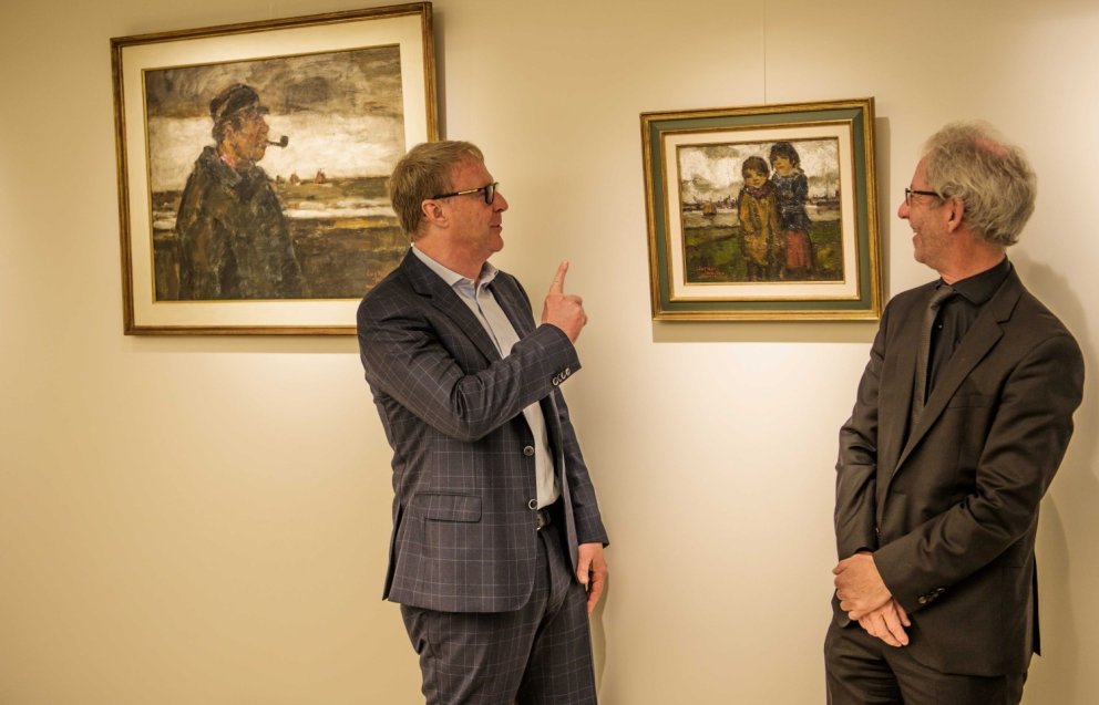 Twee mannen staan in een galerie naast schilderijen; de man links wijst naar een schilderij van twee kinderen, terwijl de rechterman naar hem kijkt.